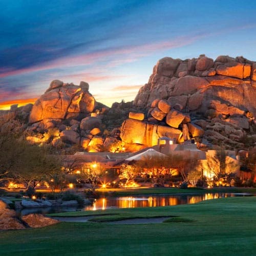 A tranquil resort with illuminated buildings at twilight, nestled among distinctive rock formations beside a calm lake in Carefree Arizona.