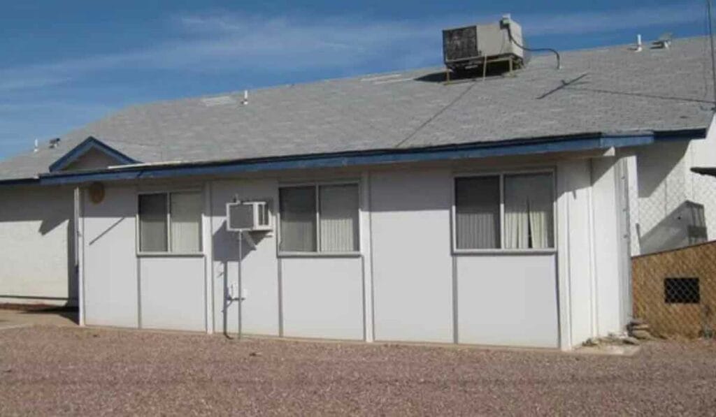 A single-story house with a grey shingle roof, an air conditioning unit, and a gravel yard under a clear blue sky.