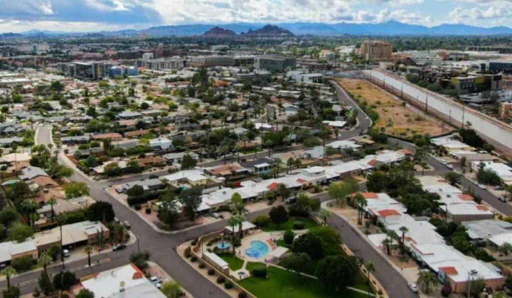 Aerial view of a suburban neighborhood with houses, greenery, and a winding road. Mountains and clouds are visible in the distance.