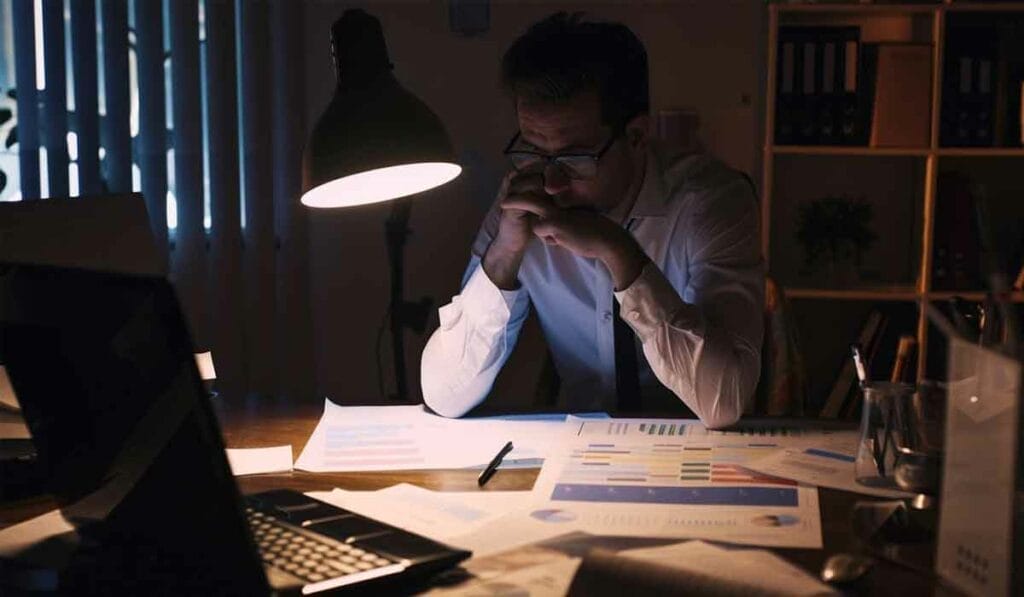 A man sits at a desk in a dimly lit room, focusing on documents and charts illuminated by a desk lamp. A laptop and more papers are scattered across the desk.