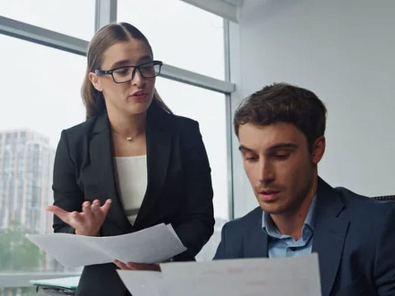 A woman in glasses stands and gestures while talking to a seated man who is looking at papers in an office setting.