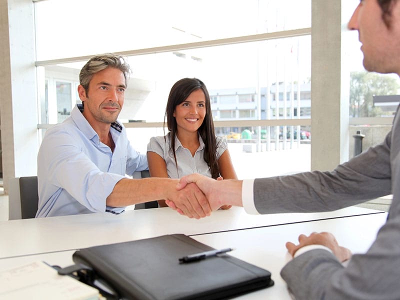 Three people in a meeting room. Two men are shaking hands across a table, while a woman sits beside them smiling. A closed notebook and pen are on the table.