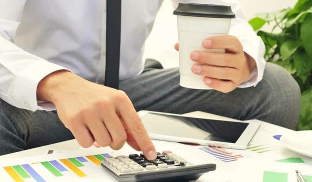 A person in business attire uses a calculator while holding a coffee cup. Nearby are a tablet and documents with charts.