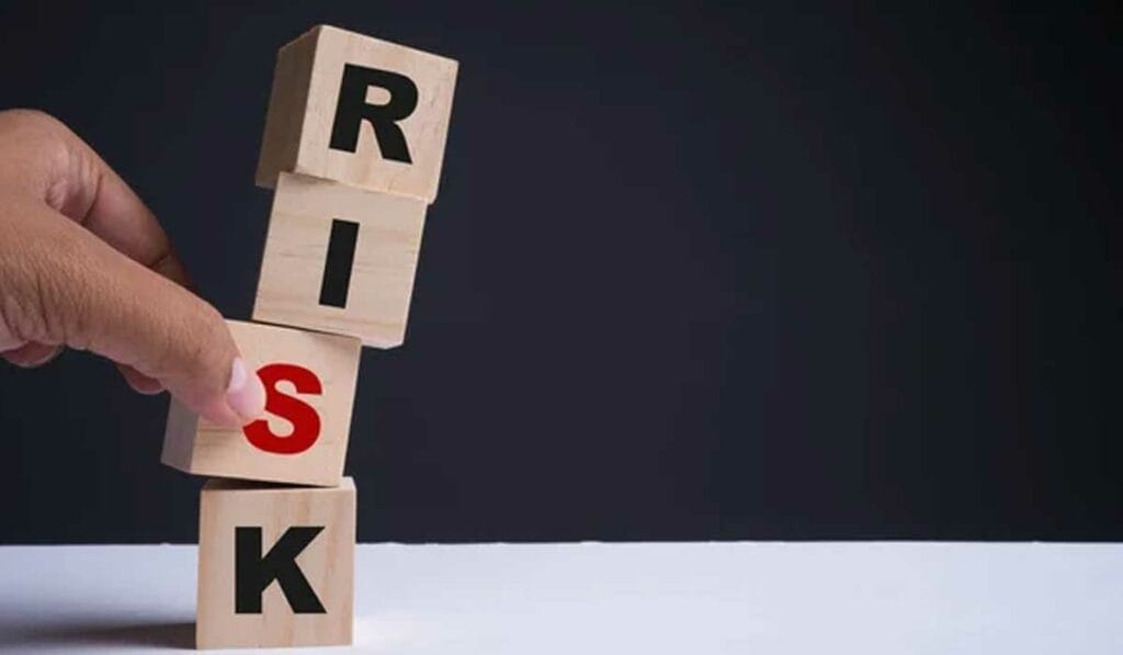 A hand attempts to stack wooden blocks spelling "RISK" vertically on a white surface against a dark background.