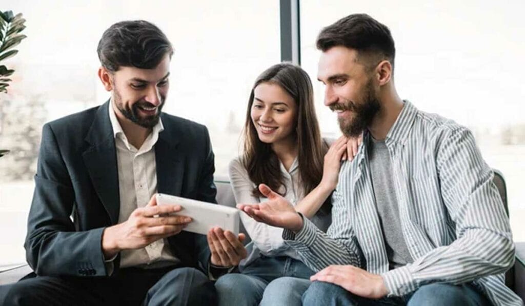 Three people sitting on a couch, engaged in discussion while looking at a tablet. Two men and one woman are smiling, wearing casual and business attire. Large window in the background.