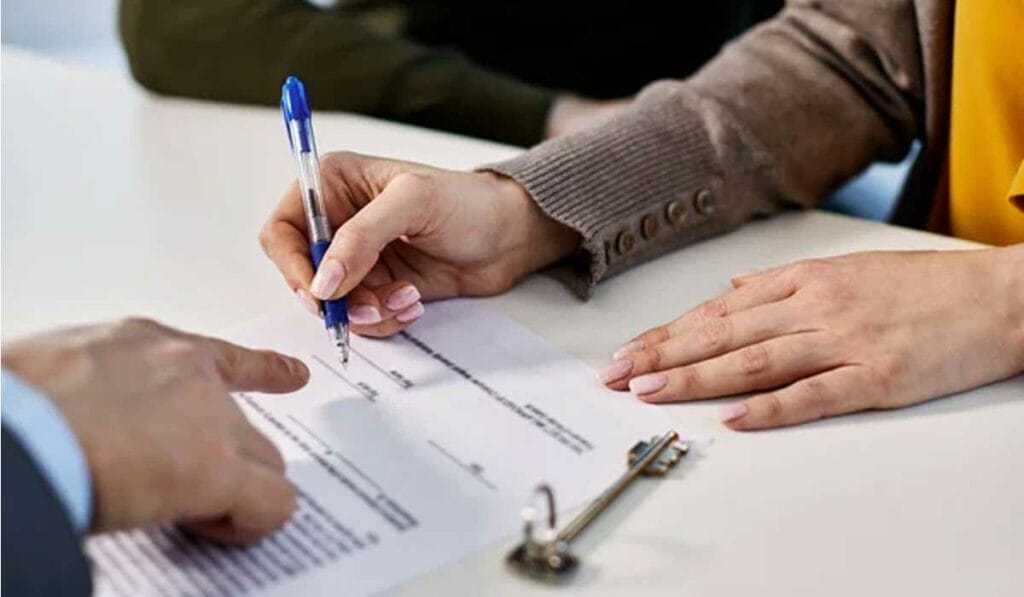 Close-up of hands signing a document; one person holds a pen while another points to the paper on a table.