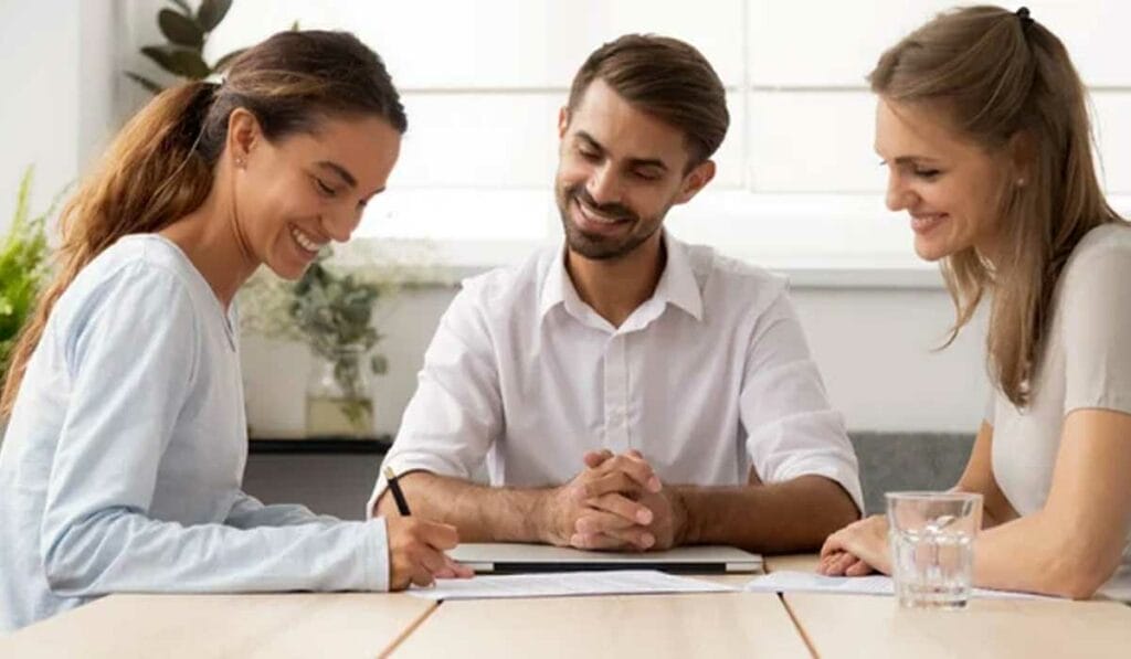 Three people are meeting at a table, with two women and one man smiling as one woman signs a document.