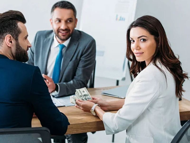 Three professionals in a meeting room, with a woman in a white blazer holding a stack of cash, and two men in suits engaged in conversation. A flip chart is visible in the background.