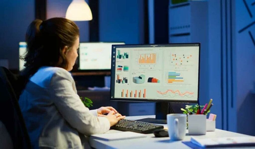 A woman sits at a desk in a dimly lit office, analyzing graphs and charts on a large computer monitor.