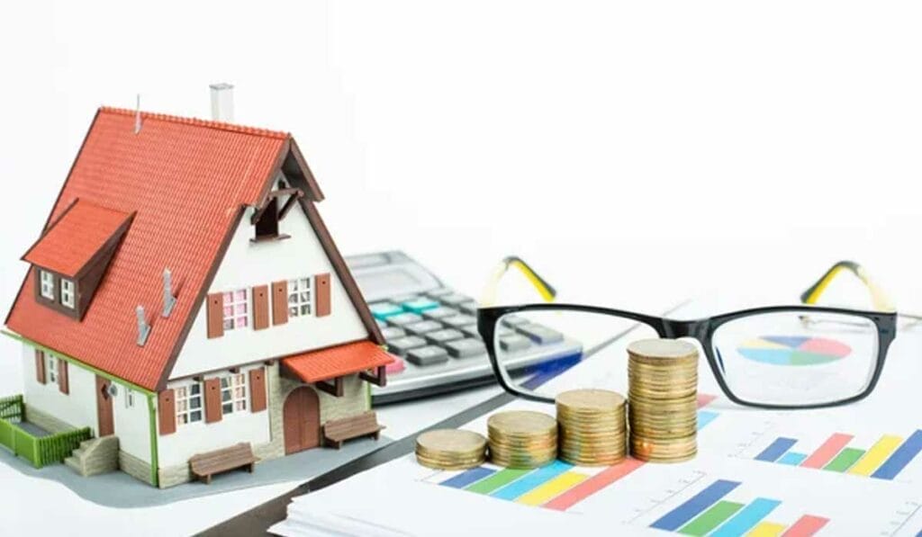 A model house, a calculator, stacks of coins, glasses, and financial charts are arranged on a table.